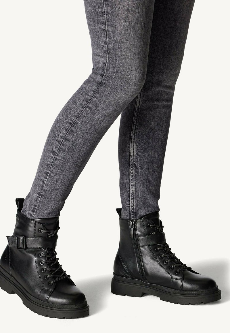 Bottes de combat en cuir noir avec lacets et fermeture éclair sur le côté, semelle en caoutchouc et détail de sangle à la cheville, associées à un jean gris ajusté.