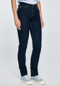 Isik, kes kannab tumel siniseid kitsaid teksaseid ja helesiniseid kõrge säärega tenniseid, seisab ühevärvilise helehalli tausta ees.