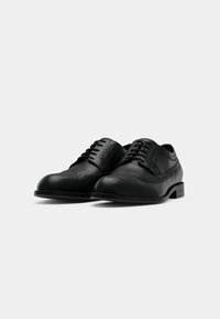 Paire de chaussures habillées brogues en cuir noir poli avec détails à bout ailé et lacets noirs sur fond blanc.