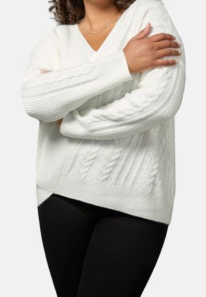 Femme croisant les bras sur la poitrine, portant un pull en maille torsadée blanc et un pantalon noir sur un fond uni.