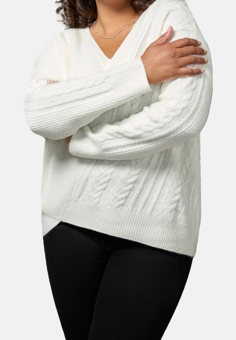 Femme croisant les bras sur la poitrine, portant un pull en maille torsadée blanc et un pantalon noir sur un fond uni.