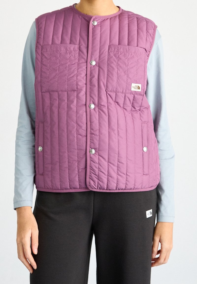 Gilet trapuntato viola con due tasche anteriori, bottoni a pressione argentati e etichetta con logo. Indossato sopra una maglietta a maniche lunghe azzurra e abbinato a pantaloni scuri.