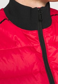 Giacca imbottita rossa con alto colletto nero, caratterizzata da una zip a tutta lunghezza e una finitura testurizzata. Dettagli neri lungo la zip.