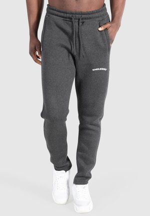 Mann trägt dunkelgraue Jogginghose mit "SMILODOX"-Logo und weißen Sneakers, eine Hand in der Tasche, vor einfachem hellem Hintergrund.