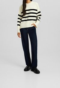 Donna che indossa un maglione crema con strisce orizzontali nere, jeans blu scuro e scarpe nere lucide, in piedi con una mano nella tasca.