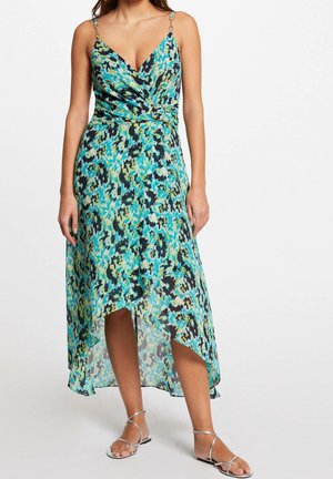 Frau trägt ein ärmelloses Wickelkleid mit V-Ausschnitt, blau-grün-schwarzem abstraktem Muster, asymmetrischem Saum und silbernen flachen Riemchensandalen.