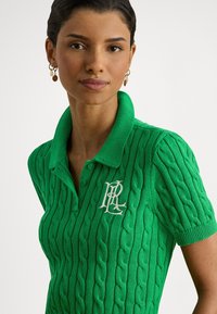 Grønn kabelstrikket poloskjorte med klassisk krage, korte ermer og en brodert hvit logo på venstre bryst.