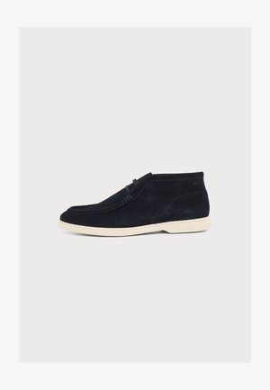 Chaussure décontractée mi-montante en suède noire avec semelle blanche et couture style mocassin, vue de côté sur fond uni.
