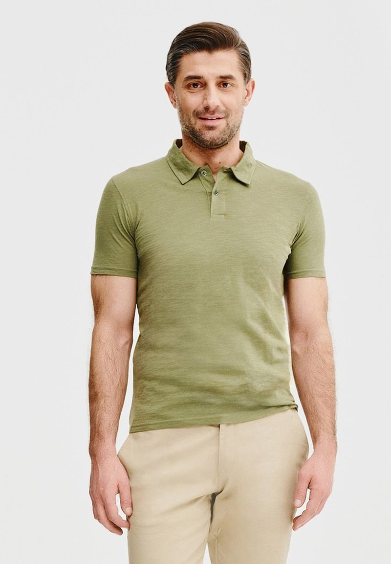 Oliwkowa koszulka polo z kołnierzykiem z przodu, krótkimi rękawami i zapięciem na trzy guziki; noszona z jasnobeżowymi spodniami. Gładki, lekki materiał.