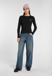 Woman wearing a light pink knit beanie, black long-sleeve fitted top, wide-leg blue jeans, and black shoes, standing with one hand in pocket.