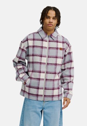 Jeune homme portant une veste zippée à carreaux violets et blancs et un jean bleu clair, debout avec une main dans sa poche.