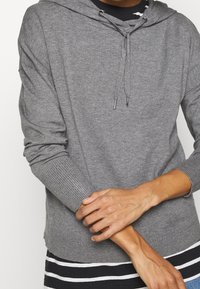 Un sweat à capuche gris avec cordon de serrage, manches longues, poignets côtelés et finition texturée, superposé sur une chemise rayée noire et blanche.