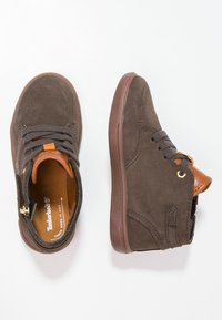 Zapatillas altas de ante marrón con cordones oscuros, forro interior naranja y suelas de goma marrón, mostradas desde vistas superiores y laterales sobre un fondo blanco.