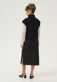 Črn pleten brezrokavnik z visokim ovratnikom, složen nad prosojno belo bluzo, v kombinaciji z oprijetim črnim krilom z bočnim razporkom in črnimi petkami.