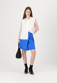 Witte mouwloze blouse met een voorknopsluiting, gecombineerd met blauwe shorts en zwarte enkelsokken. Het model houdt een gestructureerde zwarte tas vast.