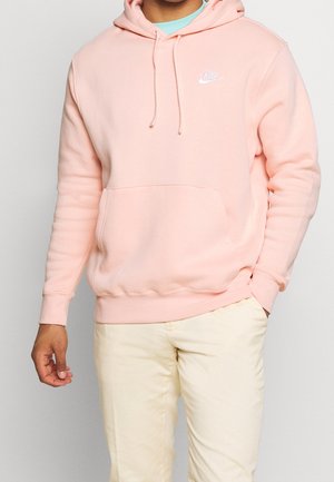 Personne portant un sweat à capuche Nike rose clair avec poche avant et cordons, associé à un pantalon blanc cassé, debout devant un fond uni.