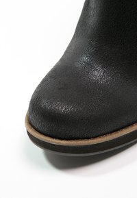 Gros plan de l'embout d'une botte en cuir noir texturé avec une bordure marron clair et une semelle noire sur fond blanc.