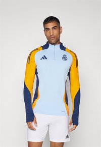 Világoskék sportos felső tengerészkék és sárga részletekkel, cipzáros gallérral, hosszú ujjakkal, és a Real Madrid logóval a mellkasán. Nedvszívó anyagból készült.