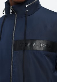 Mørkeblå jakke med høy krage, med en svart lær horisontal stripe med preget tekst. Sølvfarget glidelås og snor detaljer.