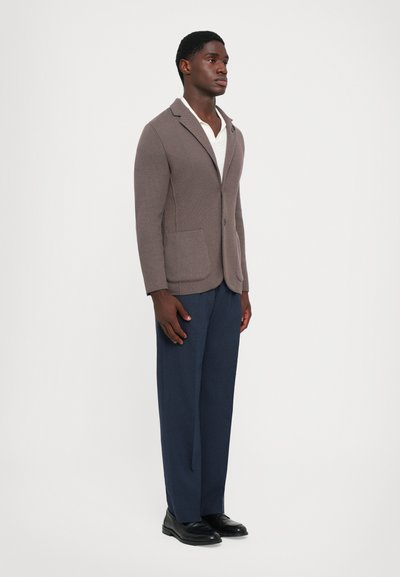 Brown knit blazer with notch lapels, two front pockets; paired with navy trousers and black shoes. White shirt underneath.