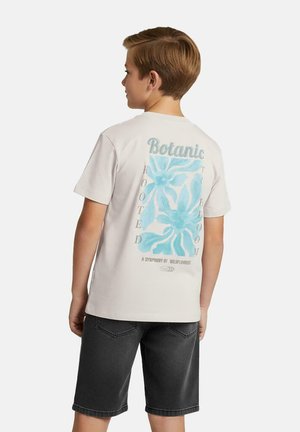 Jongen in zijaanzicht, gekleed in een wit T-shirt met blauwe bloemenprint en tekst, gecombineerd met zwarte denim shorts, tegen een effen achtergrond.