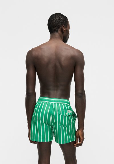 Άνδρας που στέκεται με την πλάτη, φοράει πράσινο μαγιό με κάθετες άσπρες ρίγες και πίσω τσέπη σε απλό ανοιχτό φόντο.