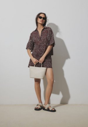 Mujer con un conjunto marrón de top y shorts con estampado floral, sandalias beige, gafas de sol negras y que sostiene un bolso tejido blanco.