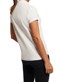 Polo blanc à manches courtes avec un col classique, présentant une texture lisse et un ourlet droit, porté avec un pantalon foncé.