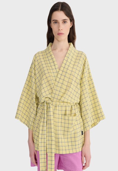 Femme portant une veste portefeuille à carreaux jaune avec des manches larges, une ceinture nouée à la taille, et un pantalon rose sur un fond clair uni.