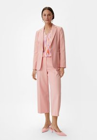 Roze maatpak met een blazer met inslagrevers en cropped wijde broekspijpen, gecombineerd met een blouse met een meerkleurig patroon en roze slingback hakken.