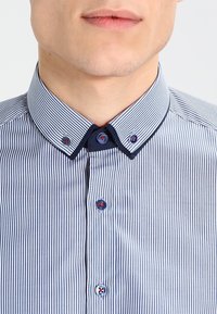 Modro-bíle pruhované tričko s kontrastním tmavě modrým límečkem, zdobené dekorativními knoflíky s červenými akcenty na límečku a manžetách.