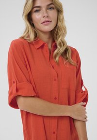Chemise rouge à boutons, avec manches retroussées, en tissu texturé, et une poche poitrine unique. Dispose de boutons assortis et d'un col classique.