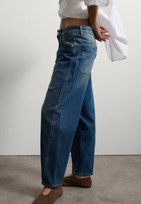 Vaqueros de denim de corte ancho en color azul, con una cintura alta, piernas rectas y un desvanecimiento sutil en la parte delantera. Combinados con una blusa blanca y zapatos marrones.