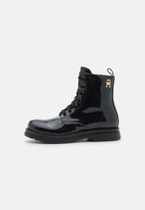 Bottine noire vernissée à lacets, avec des paillettes discrètes, une semelle épaisse et un logo TH doré sur le côté, présentée sur un fond clair uni.