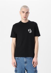 Black short-sleeve t-shirt made of cotton. Features a white graphic logo on the left chest and a round neckline. Styled with blue denim shorts.