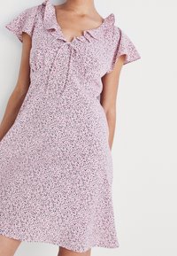 Kort rosa klänning med volangärmar, prydd med ett spritt blommönster i svart och blått. Knytning vid halsringningen.