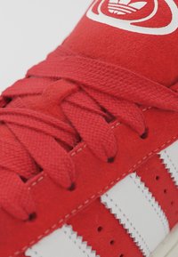 Röd mockasko med vita läderstripes, med texturerade snören och ett logopatch på tungan, som visar detaljerad sömnad.