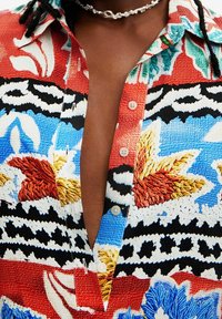 Chemise boutonnée colorée en tissu texturé avec des motifs floraux audacieux en rouge, bleu, blanc et noir. Coupe en col en V notable.