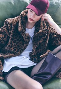 Leopardprint faux fur frakke med stor krave, parret med en hvid T-shirt og sorte shorts. Burgunder kasket og glansfuld burgunder støvle op til låret.