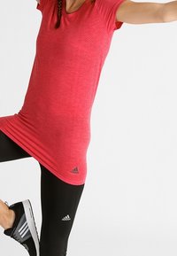 Červené sportovní tričko s krátkým rukávem, texturovaná tkanina a jemný vzor. V kombinaci s černými legínami a teniskami Adidas.