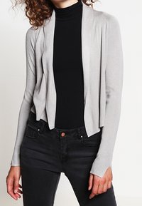 Světle šedý kardigan s otevřeným předním dílem vyrobený z měkkého pleteného materiálu, s dlouhými rukávy a uvolněným střihem, nošený přes černý top.