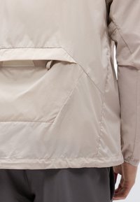 Veste beige vue de dos avec rabat de poche angulaire, surpiqûres visibles et ourlet élastiqué, portée sur un pantalon foncé.