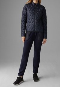 Tmavě modrá prošívaná bunda s vysokým límcem, se zapínáním na přední zip a dlouhými rukávy, kombinovaná s tmavě modrými tepláky a sportovními botami.