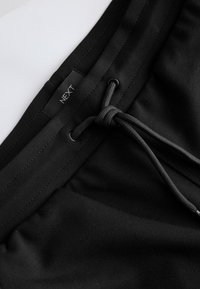 Juodi šortai su tekstūruota medžiaga, turintys reguliuojamą raištelį ir etiketę su užrašu „NEXT“ netoli juosmens.
