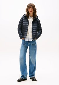 Ženska stoji z rokami v žepih, oblečena v črno puhasto jakno, belo majico, kavbojke in črne čevlje na belem ozadju.