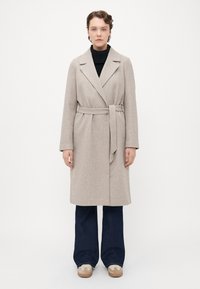 Kvinne står iført en beige belagt ullfrakk over en svart topp, mørke vide bukser og beige joggesko mot en ensfarget hvit bakgrunn.