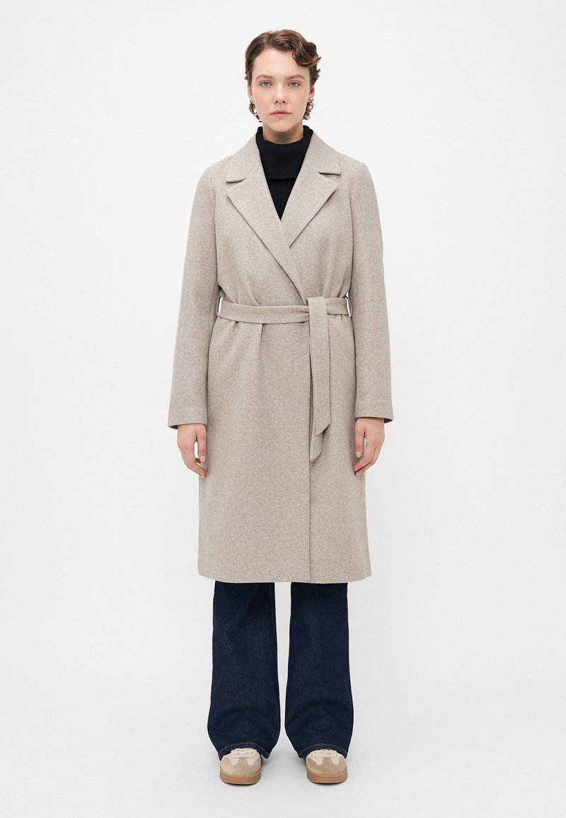 Kvinne står iført en beige belagt ullfrakk over en svart topp, mørke vide bukser og beige joggesko mot en ensfarget hvit bakgrunn.