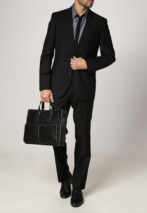 Černý na míru šitý oblek s klopou s výstřihem, šedá košile s kravatou a strukturovaná černá kožená aktovka se dvěma předními kapsami.