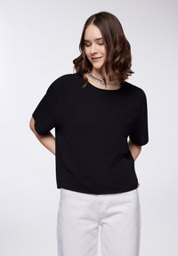 Top corto nero a maniche corte con una vestibilità rilassata e tessuto texturizzato. Indossato con pantaloni bianchi e una collana scintillante. Design semplice e moderno.