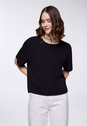 Top corto nero a maniche corte con una vestibilità rilassata e tessuto texturizzato. Indossato con pantaloni bianchi e una collana scintillante. Design semplice e moderno.